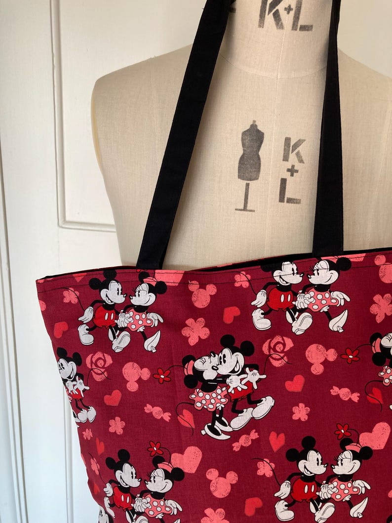 May include: A red tote bag with a black handle. The bag is decorated with a repeating pattern of Mickey Mouse and Minnie Mouse in a romantic pose. The pattern also includes hearts and roses.