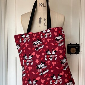 May include: A red tote bag with a repeating pattern of Mickey Mouse and Minnie Mouse holding hands. The pattern also includes pink hearts and flowers. The bag has black straps.