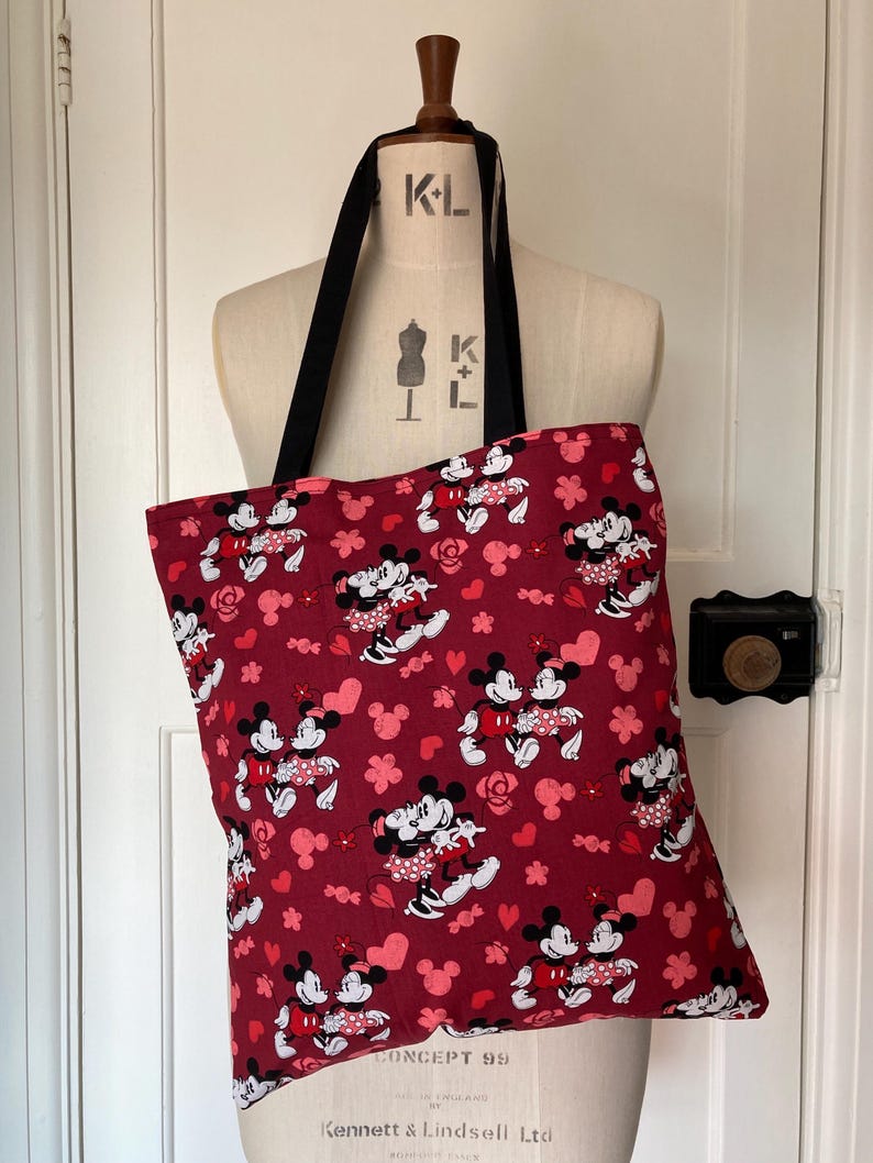 May include: A red tote bag with a repeating pattern of Mickey Mouse and Minnie Mouse holding hands. The pattern also includes pink hearts and flowers. The bag has black straps.