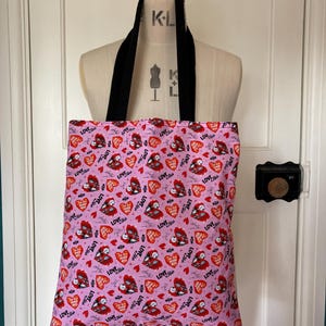 May include: A pink tote bag with black straps, featuring a repeating pattern of red hearts, bats, and the word "LOVE" in a handwritten style. The bag is displayed on a mannequin.