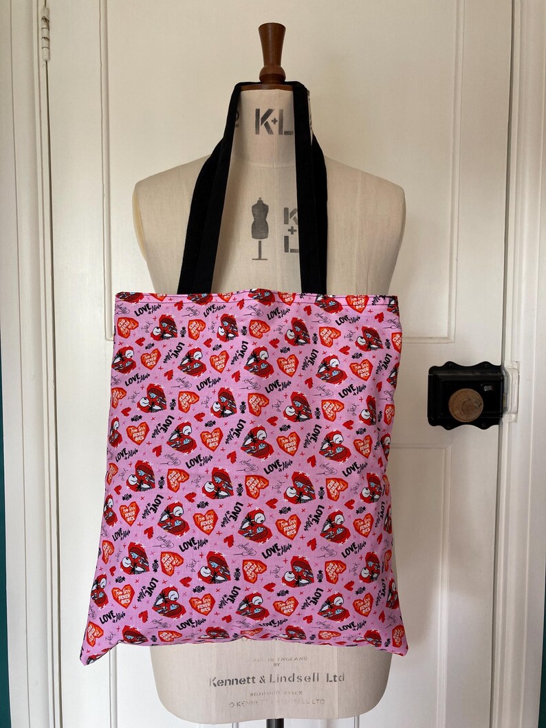 May include: A pink tote bag with black straps, featuring a repeating pattern of red hearts, bats, and the word "LOVE" in a handwritten style. The bag is displayed on a mannequin.