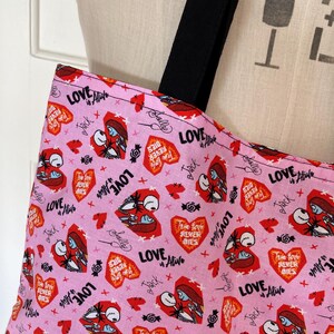 May include: Pink tote bag with black straps featuring a repeating pattern of red hearts, Jack and Sally illustrations, and the phrases "Love Alive" and "True Love Never Dies." The bag is made of fabric.