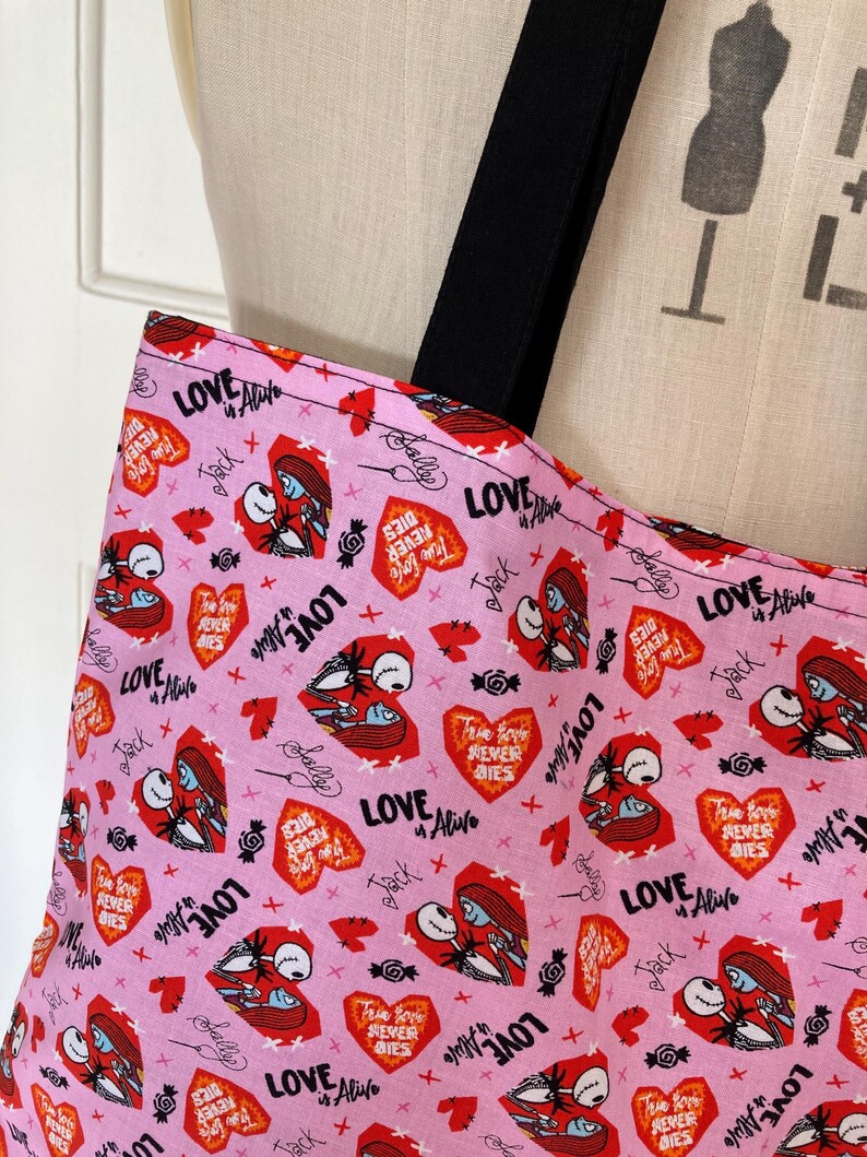 May include: Pink tote bag with black straps featuring a repeating pattern of red hearts, Jack and Sally illustrations, and the phrases "Love Alive" and "True Love Never Dies." The bag is made of fabric.