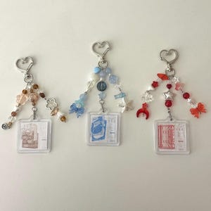 May include: Three beaded keychains with heart-shaped clasps. Each keychain has a clear plastic tag with a printed design. The beads are in shades of brown, blue, and red, with decorative charms like flowers, stars, and bows.