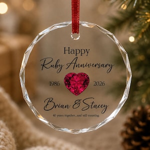 May include: A round, faceted clear glass ornament hanging from a red ribbon. The text reads Happy Ruby Anniversary, 1986, 2026, Brian & Stacey, and 40 years together...and still counting. A vibrant red heart-shaped gemstone is centered on the glass.