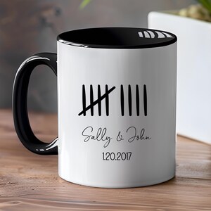 Puede incluir: Taza de cerámica blanca con interior, asa y borde negros. La taza presenta un diseño de marcas de conteo con los nombres "Sally & John" y la fecha "1.20.2017" impresos en negro.
