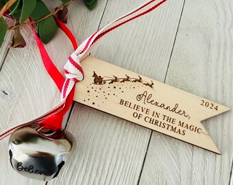 May include: A wooden Christmas ornament with the name "Alexander" and the year "2024" engraved. The ornament features a sleigh and reindeer design, with the text "Believe in the Magic of Christmas." A silver bell and red and white striped ribbon are attached.