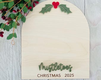 May include: An arched light-colored wooden sign featuring a decorative green holly leaf and red berry accent at the top. The sign displays the text Mistletoes in a raised green script font above the printed text CHRISTMAS 2025.