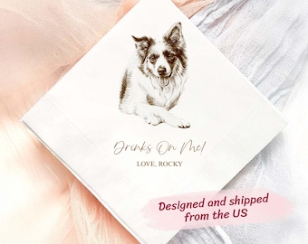 May include: White square cocktail napkin featuring a detailed illustration of a dog with black and white markings. The text "Drinks On Me! Love, Rocky" is printed below the dog image. The napkin is set against a soft, textured background.