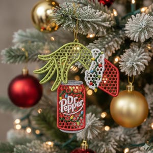 May include: Embroidered Christmas ornament featuring a green hand holding a red Dr. Pepper can with a string of lights. The ornament also includes a red and white stocking with a string of lights. The ornament is hanging on a Christmas tree.