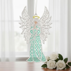 Può includere: Un ornamento a forma di angelo ricamato bianco con ali dettagliate e un vestito verde menta. L'angelo ha un'aureola dorata ed è sospeso da un filo d'oro. L'ornamento è esposto davanti a una finestra con rose bianche.