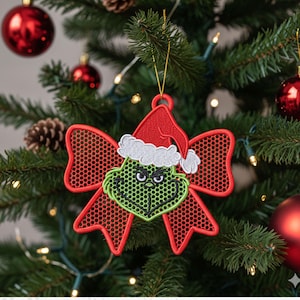 May include: A Christmas tree decoration featuring a green Grinch face wearing a red Santa hat with white trim. The Grinch is set against a red bow-shaped background with a mesh design. The ornament hangs from a gold string on a Christmas tree.