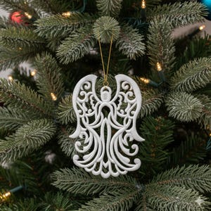 May include: White angel Christmas ornament with intricate cut-out design, hanging on a green Christmas tree. The ornament has a gold string for hanging. The tree is decorated with small lights.