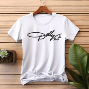 May include: A white short-sleeved t-shirt featuring the black cursive text Dolly followed by XOXO and a small heart symbol printed on the center of the chest. The garment has a round neckline and is displayed on a wooden hanger against a slatted background.