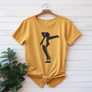 May include: Mustard yellow t-shirt with a black silhouette of a dancer in a famous pose. The t-shirt is knotted at the bottom and hangs on a wooden hanger against a white wood-panelled wall.