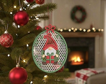 May include: An oval-shaped Christmas ornament featuring an embroidered gnome wearing a red hat and holding a green gift. The gnome is centered within a green frame with a white honeycomb pattern background and a red bow at the top for hanging.