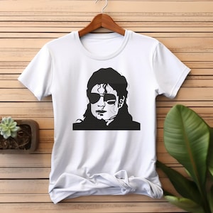 May include: White t-shirt with a black and white graphic portrait of a person wearing sunglasses. The t-shirt is displayed on a wooden hanger. The background is wooden.