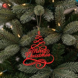 May include: A red, script-style Christmas tree ornament featuring the text Merry Christmas in a continuous, swirling line design. The ornament is suspended by a thin gold string and hangs against the green needles of a pine tree.