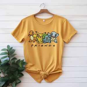 Könnte beinhalten: Senfgelbes T-Shirt mit einer Grafik von Zeichentrickfiguren und dem Wort "FRIENDS". Die Figuren sind in Orange, Gelb, Grün und Blau gehalten. Das T-Shirt hängt an einem Holzbügel vor einem weißen Holzgrund.