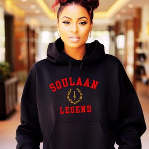 Puede incluir: Sudadera con capucha negra con el texto "SOULAAN LEGEND" en rojo, con un emblema dorado. La sudadera tiene un bolsillo delantero y una capucha con cordón. La modelo tiene el pelo rojo y lleva pendientes de aro.