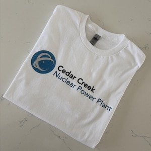 May include: White t-shirt with a blue and gray logo and text that reads "Cedar Creek Nuclear Power Plant". The shirt is folded neatly on a light-colored surface.