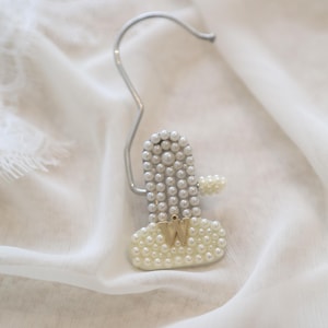 May include: A cactus-shaped decorative clip adorned with white faux pearls. The vertical column and horizontal arm are covered in pearls, while the base features a gold-toned letter W. A silver-colored metal hook is attached to the top of the clip.