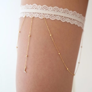 May include: A white lace garter featuring a delicate gold-toned chain fringe. The thin gold chains are accented with small metallic beads and a single dangling letter L charm encrusted with clear stones, creating a layered accessory design.