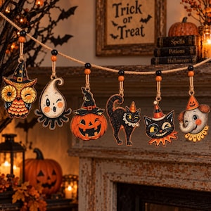 May include: A Halloween-themed garland featuring six hanging wooden ornaments suspended from a jute rope with black and orange beads. The ornaments depict an owl, a ghost, a jack-o'-lantern, a black cat, a cat face, and an elephant, all wearing hats.