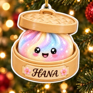Könnte beinhalten: Ein hängendes Ornament in Form eines Bambusdampfkorbs, das ein glitzerndes, regenbogenfarbenes Bao-Brötchen mit einem lächelnden Gesicht enthält. Vorne am Dampfkorb befindet sich ein rechteckiges Schild mit dem Namen HANA und zwei kleinen rosa Kirschblüten.