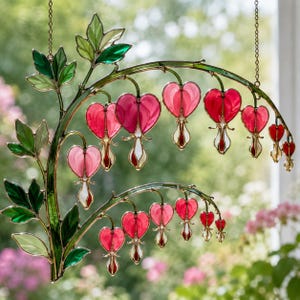 Puede incluir: Un atrapasoles de vidrieras con un diseño de planta de corazón sangrante con dos tallos curvos. La pieza muestra múltiples flores en forma de corazón en tonos rosa y rojo con acentos blancos, complementadas con hojas de vidrio verde y soldadura.