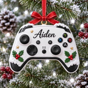 May include: A white video game controller ornament featuring the name Aiden in black script, accented with green holly leaves, red berries, small gold stars, and white snow-like trim, topped with a red ribbon bow for hanging on a tree.