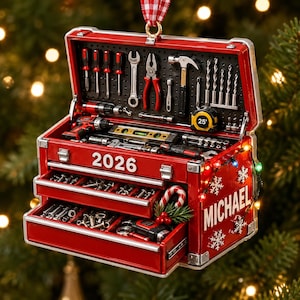 May include: A red toolbox ornament featuring a pegboard interior with miniature tools, a tape measure, and drill bits. The exterior displays the year 2026, the name MICHAEL, white snowflake accents, and a small candy cane decoration with greenery.