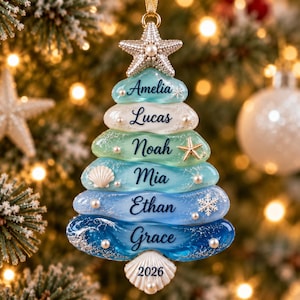 May include: A Christmas tree-shaped ornament made of stacked, smooth, sea-glass-coloured layers in shades of teal and blue. Each layer features a printed name: Amelia, Lucas, Noah, Mia, Ethan, and Grace. It is topped with a starfish and includes small pearl accents, a seashell, and a snowflake. The base is a white seashell with the year 2026 printed on it.