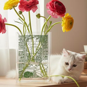 May include: A clear rectangular glass vase featuring a green line-art illustration of a library interior with the text Kendra's Library. The vase holds several vibrant red and yellow ranunculus flowers, with one stem resting near a white cat.