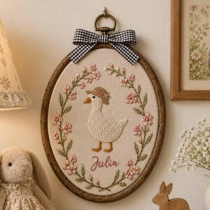May include: An oval embroidery hoop wall hanging featuring a cream fabric background with a stitched white duck wearing a floral hat, surrounded by a pink flower wreath and the name Julia in cursive script, topped with a black and white gingham bow.