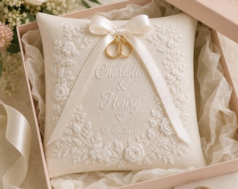 May include: A square cream-colored ring pillow featuring white floral embroidery, a satin ribbon bow, and two gold rings. The text reads Charlotte & Henry with the date 08.09.2026 and a small heart icon, all presented in a light pink box.