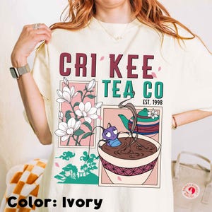 May include: Ivory-colored t-shirt featuring the text "Cri Kee Tea Co. Est. 1998." The graphic includes illustrations of flowers, a teapot, and a cartoon character in a teacup. The shirt's color is ivory.