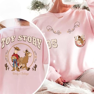 May include: A light pink sweatshirt featuring graphics from the movie "Toy Story." The back of the shirt displays "Toy Story" text above an image of Woody and Bullseye. The front has decorative swirls, the number "95," and a horseshoe with a horse's face.