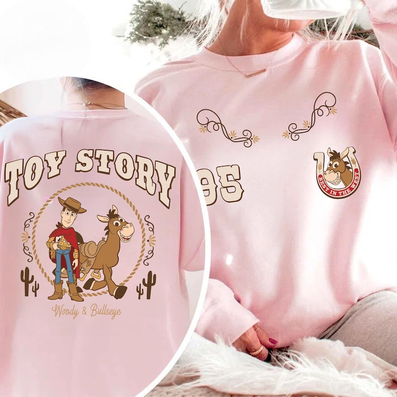 May include: A light pink sweatshirt featuring graphics from the movie "Toy Story." The back of the shirt displays "Toy Story" text above an image of Woody and Bullseye. The front has decorative swirls, the number "95," and a horseshoe with a horse's face.