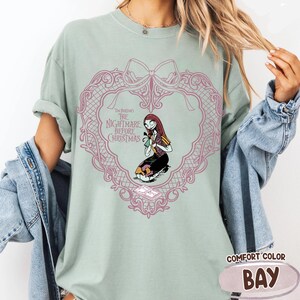 May include: Pale green t-shirt with a pink heart-shaped design and the text "The Nightmare Before Christmas." The design features Sally from the film. The shirt is a Comfort Color brand in the colour Bay.