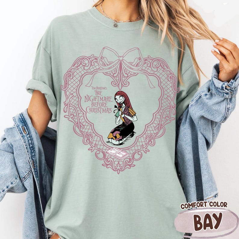 May include: Pale green t-shirt with a pink heart-shaped design and the text "The Nightmare Before Christmas." The design features Sally from the film. The shirt is a Comfort Color brand in the colour Bay.
