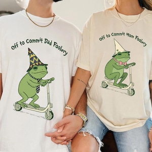 May include: Two off-white t-shirts with the text "Off to Commit Dad Foolery" and "Off to Commit Mom Foolery". Each shirt features a green frog riding a scooter and wearing a hat and tie or bow tie. The frog illustrations are in a cartoon style.