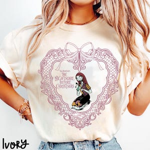 May include: Ivory t-shirt with a design from "The Nightmare Before Christmas." The design features a heart-shaped frame with Sally kneeling, holding a flower. The text "Tim Burton's The Nightmare Before Christmas" is also visible.