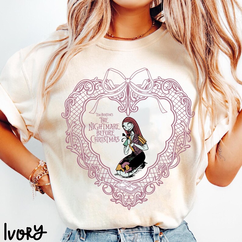 May include: Ivory t-shirt with a design from "The Nightmare Before Christmas." The design features a heart-shaped frame with Sally kneeling, holding a flower. The text "Tim Burton's The Nightmare Before Christmas" is also visible.