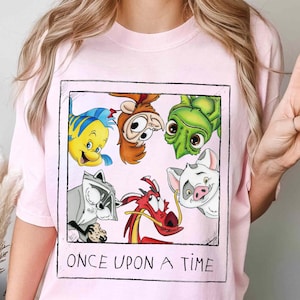 May include: Light pink t-shirt featuring a framed graphic with cartoon characters, including a yellow fish, a monkey, a green turtle, a raccoon, a red dragon, and a pig. The text "ONCE UPON A TIME" is printed below the graphic.