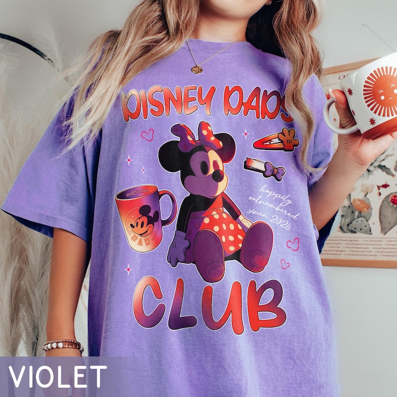 Może przedstawiać: Fioletowa koszulka z grafiką Myszki Minnie. Projekt zawiera napisy "Disney Dads Club" i "happily outnumbered since 2026". Na koszulce znajduje się r&oacute;wnież kubek do kawy z Myszka Miki.