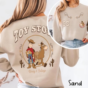 May include: Sand-colored sweatshirt featuring a graphic of Woody and Bullseye from Toy Story, encircled by a rope design. The back design includes the text "TOY STORY". The front has a smaller design with the number "95" and a horseshoe.