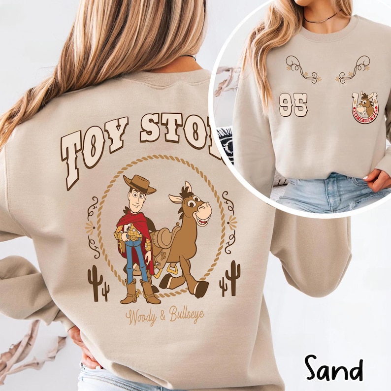 May include: Sand-colored sweatshirt featuring a graphic of Woody and Bullseye from Toy Story, encircled by a rope design. The back design includes the text "TOY STORY". The front has a smaller design with the number "95" and a horseshoe.