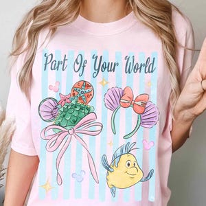 Puede incluir: Una camiseta rosa claro con el texto Part Of Your World en letra cursiva verde azulado oscuro. El gr&aacute;fico incluye una diadema de orejas con patr&oacute;n de escamas verdes y lazo rosa, una diadema de conchas moradas y un pez amarillo.