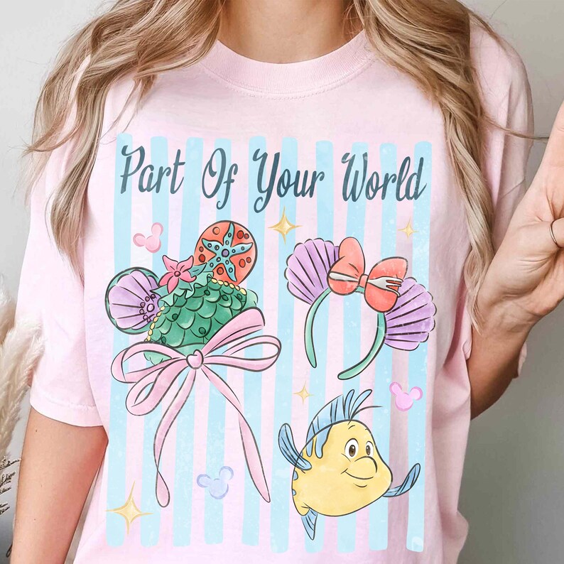 Puede incluir: Una camiseta rosa claro con el texto Part Of Your World en letra cursiva verde azulado oscuro. El gr&aacute;fico incluye una diadema de orejas con patr&oacute;n de escamas verdes y lazo rosa, una diadema de conchas moradas y un pez amarillo.